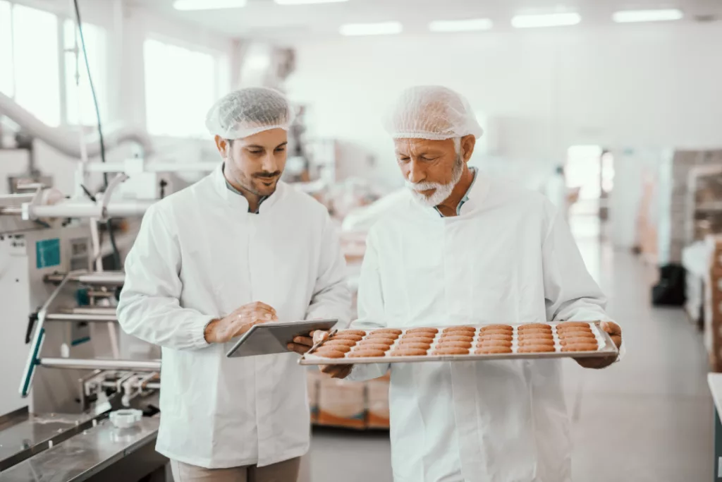 Senior employee holding tray with fresh cookies while supervisor evaluating quality using bakery erp system in a tablet. Both are dressed in sterile white uniforms and having hairnets. Food plant interior.