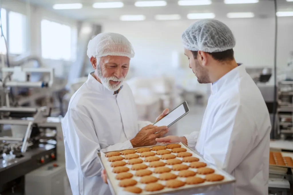 Young employee holding tray with fresh cookies while supervisor evaluating quality and holding tablet using Panni's Sage X3 ERP system. ERP-Driven Transformation in the Confectionery Industry
